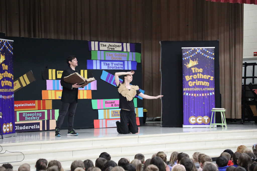 A theatrical performer in a black blazer stands on a stage holding a large, weathered open book while looking toward another performer who is kneeling and looking upward with one hand to their forehead. The backdrop features oversized colorful book spines labeled with fairy tale titles like The Juniper Tree and Sleeping Beauty, alongside a banner for The Brothers Grimm Spectacular by CCT.