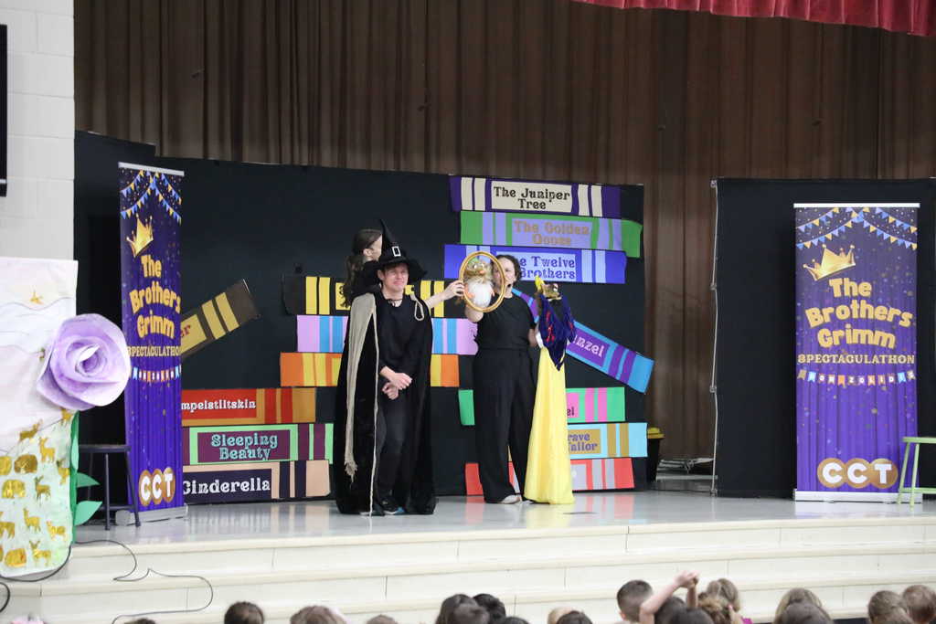 Three actors perform on a stage in front of a backdrop of oversized book spines. One actor wears a tall black witch's hat and a tan cape, holding a small puppet in a gold frame, while another actor holds up a yellow dress and a third stands behind them, all performing for a large audience of children.