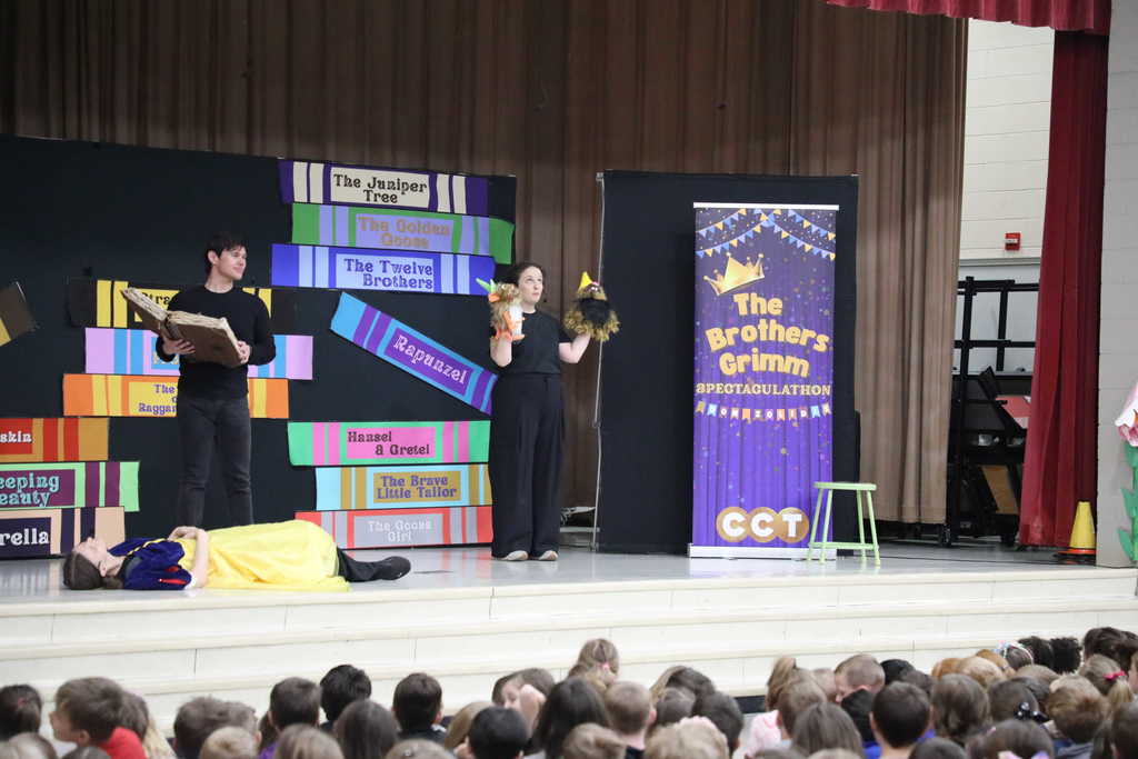 A theatrical scene on stage shows one performer lying flat on the floor in a Snow White costume while a male performer stands over them holding a large prop book. To the right, a female performer in black holds up two puppets, all set against a backdrop of giant colorful book spines and a purple Brothers Grimm banner.