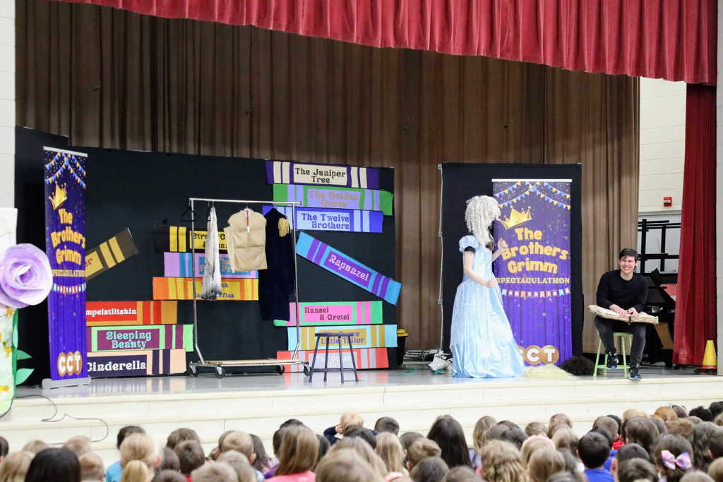 A stage setup for a play titled The Brothers Grimm Spectacular features a performer in a shimmering light blue ballgown and a large white wig walking across the stage. To the right, a male performer sits on a stool with a large prop book on his lap, and a clothing rack with various costumes stands to the left.
