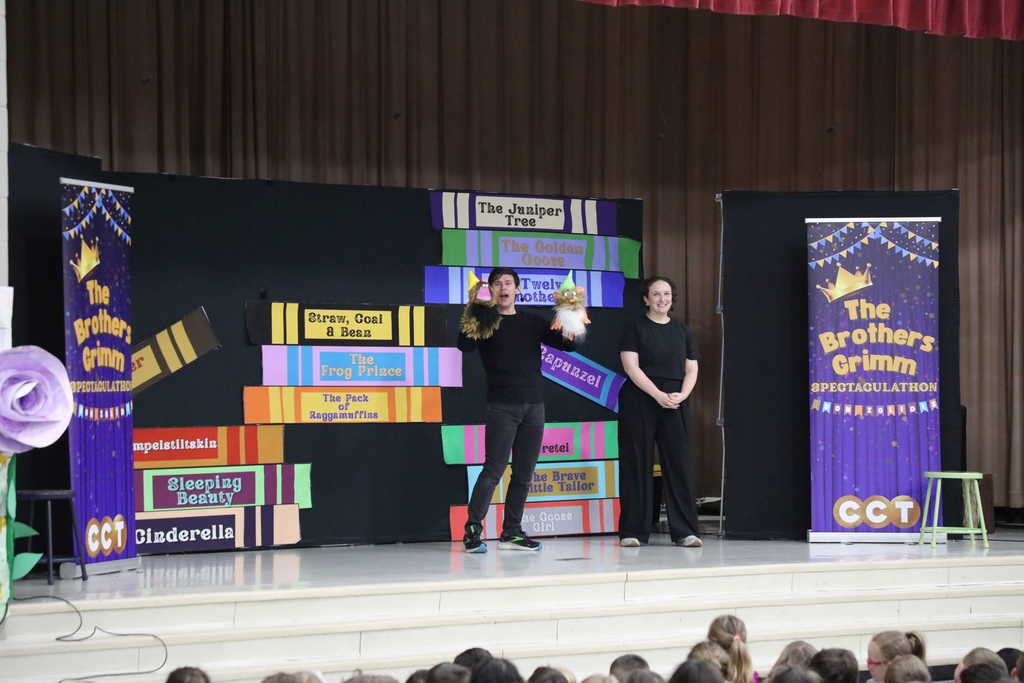 A male performer on a stage holds up two whimsical puppets with pointed yellow hats while a female performer in a black outfit stands smiling next to him. They are positioned in front of a colorful set piece designed to look like a stack of giant books, with an audience of children visible in the foreground.