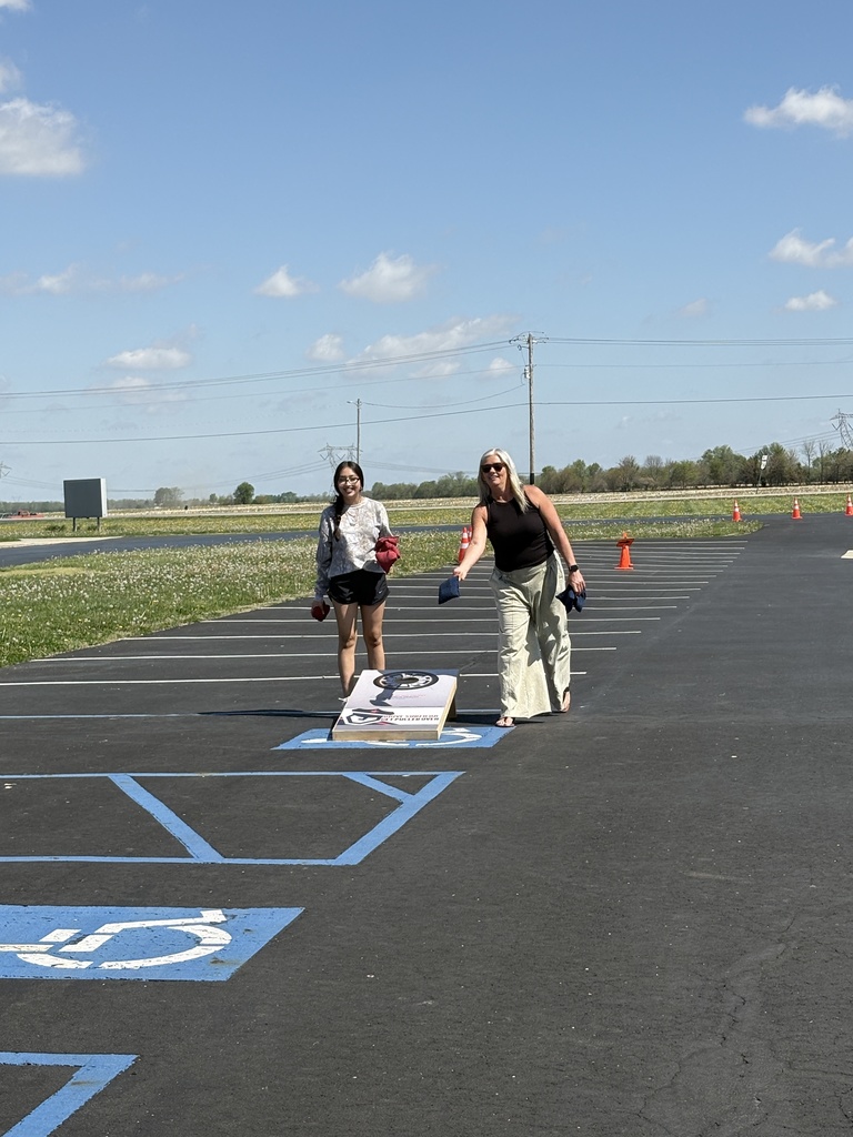 Two women play a game of cornhole in a paved parking lot under a bright blue sky, with one woman in a black tank top and khaki pants captured mid-toss.