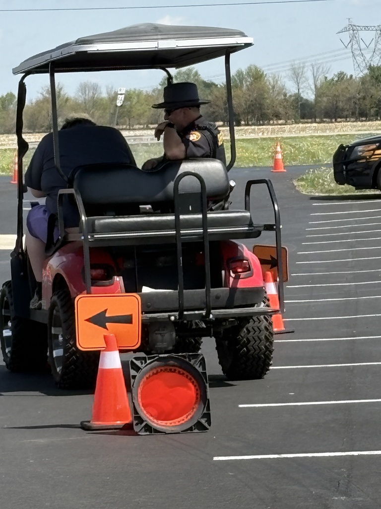 A red golf cart driven by a person in a dark shirt and purple shorts navigates a course marked with orange cones and directional signs, with a Sheriff's deputy riding in the passenger seat.