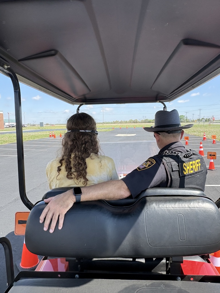 An over-the-shoulder view from the back of a red golf cart shows a young woman with long curly hair wearing impairment goggles while a Sheriff's deputy sits beside her, looking out toward a course marked with orange cones.