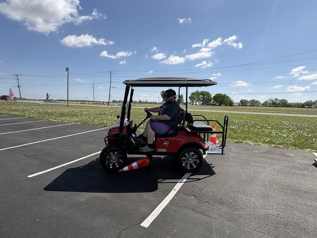 A side view shows a young man wearing black impairment goggles driving a red golf cart through a parking lot, accidentally knocking over an orange traffic cone under the front tire.