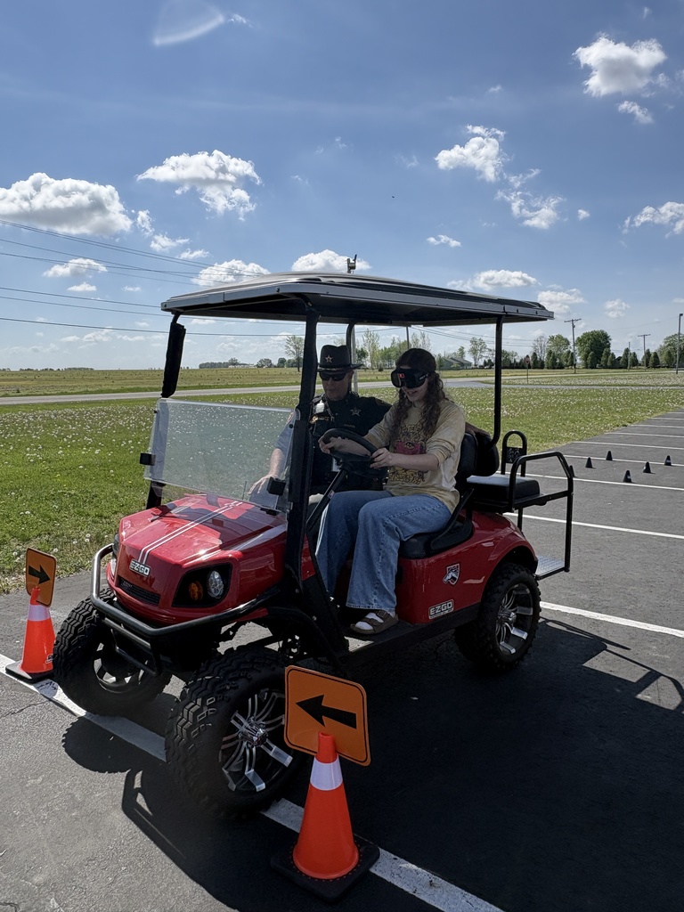 A young woman wearing impairment simulation goggles sits in the driver's seat of a red golf cart in a parking lot, accompanied by a law enforcement officer in a tan uniform and hat.