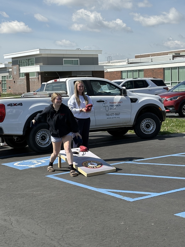 A young woman in a black hoodie and shorts tosses a beanbag toward a cornhole board decorated with a "Click It or Ticket" logo, while another woman stands behind her next to a white pickup truck.