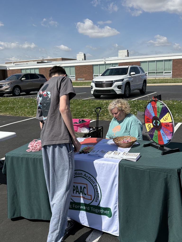 A young man in a gray t-shirt and sweatpants stands at an outdoor table covered with a green cloth featuring the Pickaway Addiction Action Coalition logo, while a woman in a teal shirt assists him behind a colorful prize wheel.