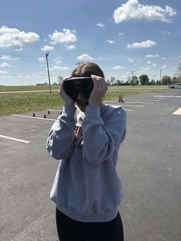 A young woman in a gray sweatshirt stands in a parking lot on a sunny day, holding a pair of black "Fatal Vision" goggles over her eyes to simulate impairment.