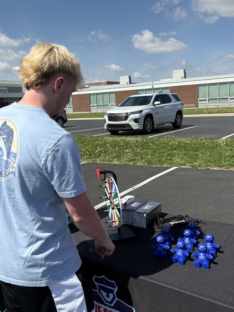 A blonde young man in a light blue t-shirt stands at a table in a parking lot, looking down at a small prize wheel and several small blue figurines shaped like people.