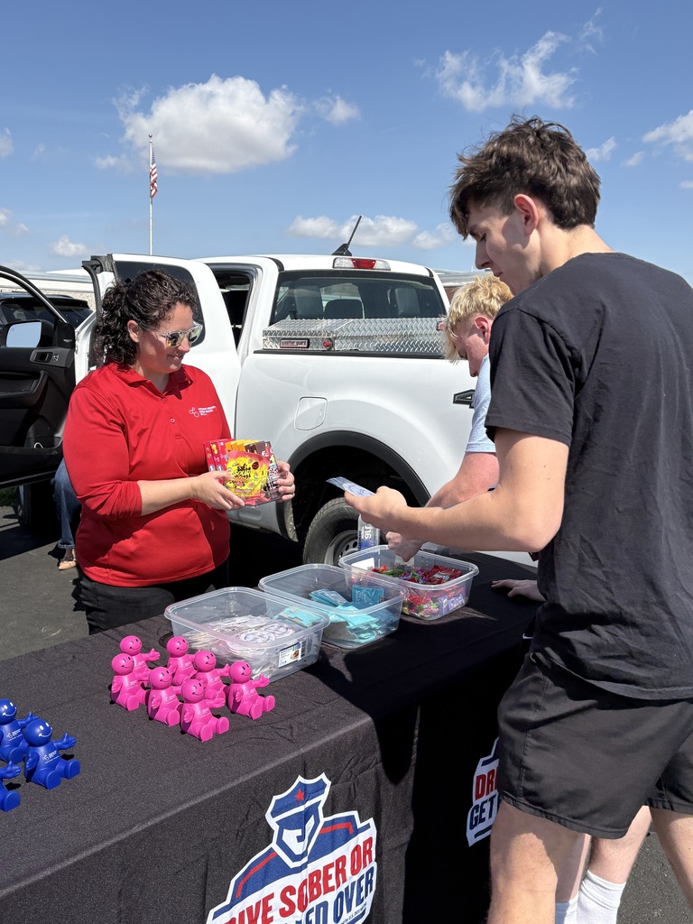 A woman in a red polo shirt stands behind a table in a parking lot, handing a colorful box of snacks to a young man while other small prizes and clear bins sit on the black tablecloth.