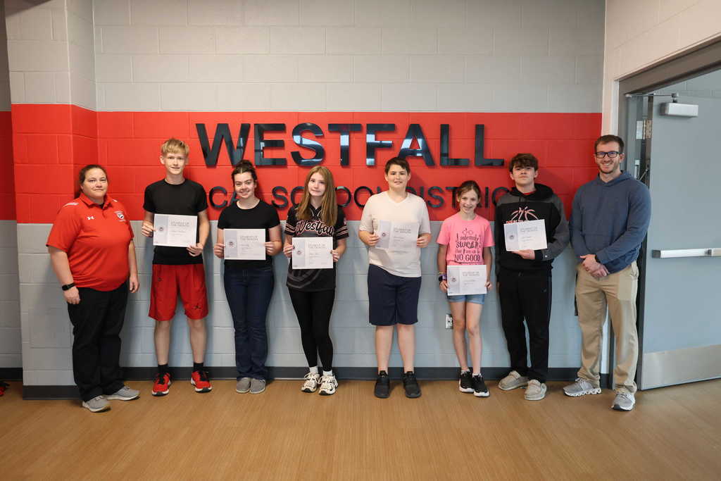 Six middle school students stand in a line holding "Student of the Month" certificates in front of a gray brick wall with a red stripe and the words "WESTFALL LOCAL SCHOOL DISTRICT" in black lettering. They are flanked by two adult staff members, one woman in a red polo shirt on the far left and one man in a blue hoodie and khaki pants on the far right. The students are dressed in casual school attire, including t-shirts, hoodies, jeans, and athletic shorts.