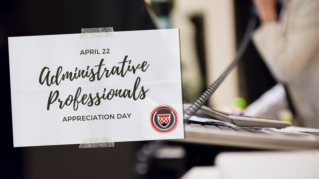 Administrative Professionals Day: A printed sign taped to a desk reads “April 22 Administrative Professionals Appreciation Day” in a mix of bold and script fonts. The sign features a red and black “W” logo in the corner. In the blurred background, a desk phone and office setting are visible, suggesting a workplace environment.