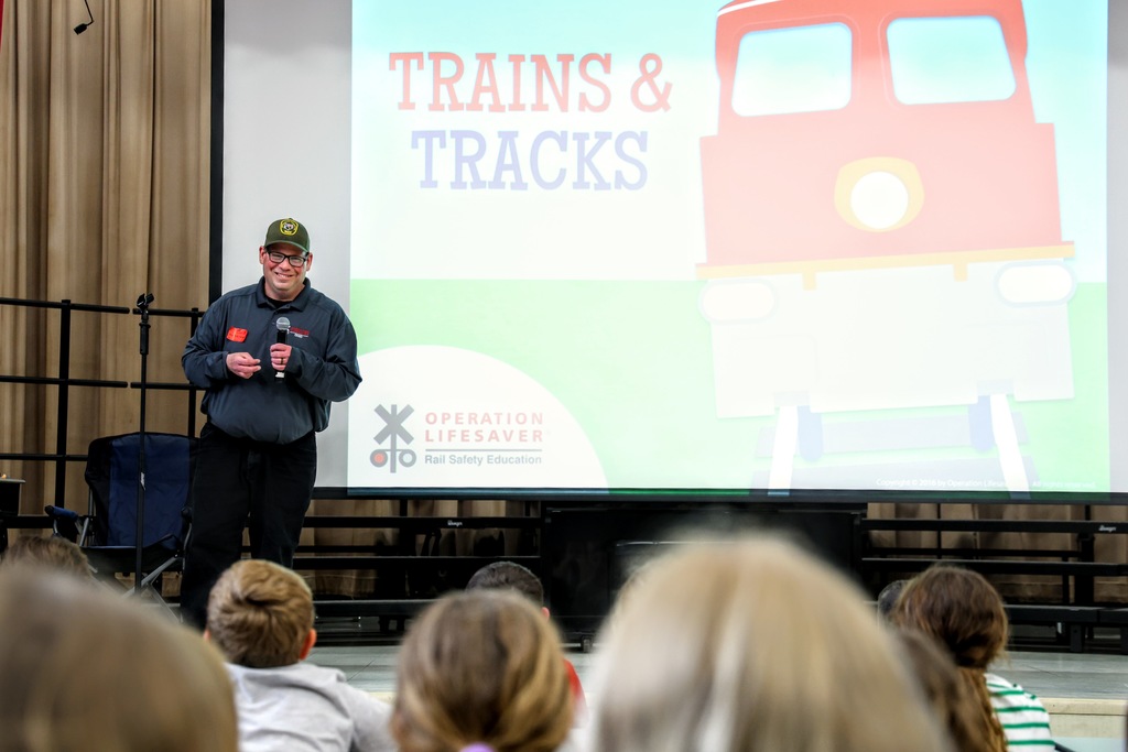 Rail Safety Inspector Joshua Elzey speaking to students in audience with a screen in the background showing Trains and Tracks with a red train engine