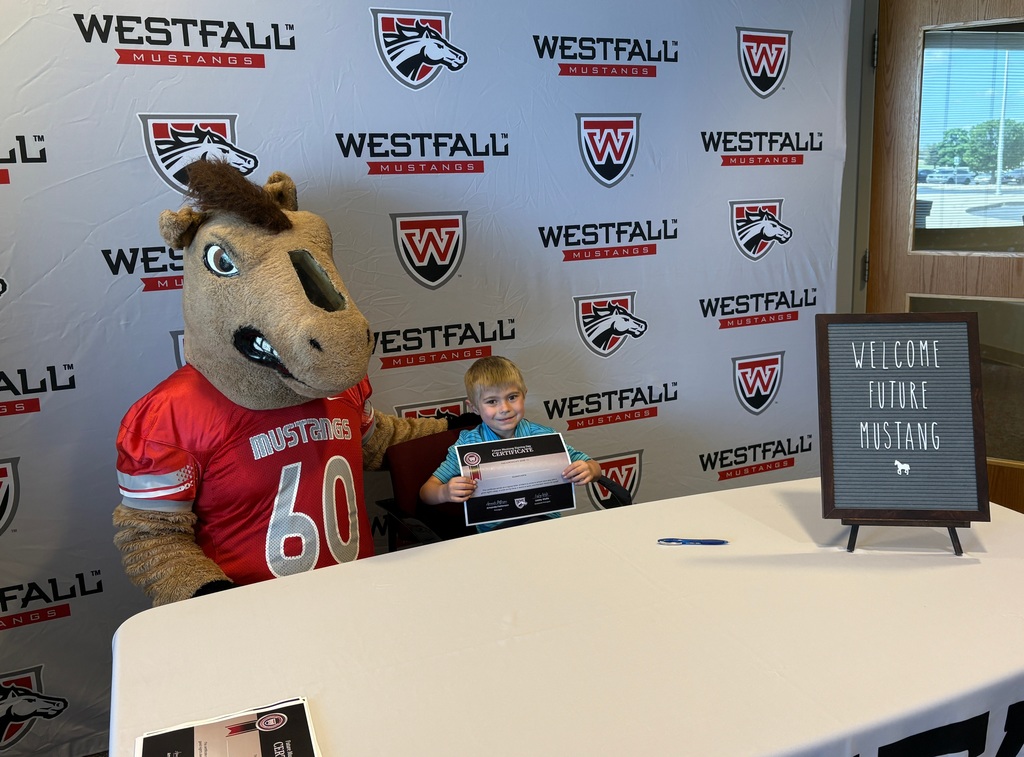 Kindergartener signing a certificate with the Westfall backdrop in the background and a sign that reads Welcome Future Mustang
