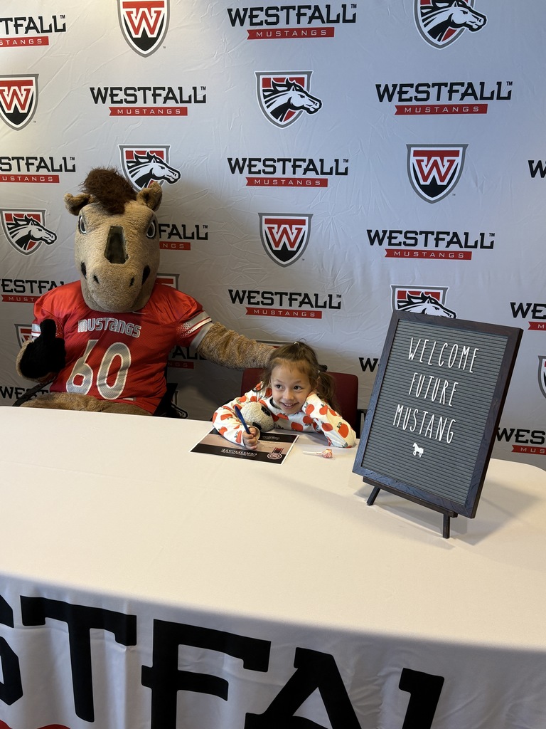 Kindergartener signing a certificate with the Westfall backdrop in the background and a sign that reads Welcome Future Mustang