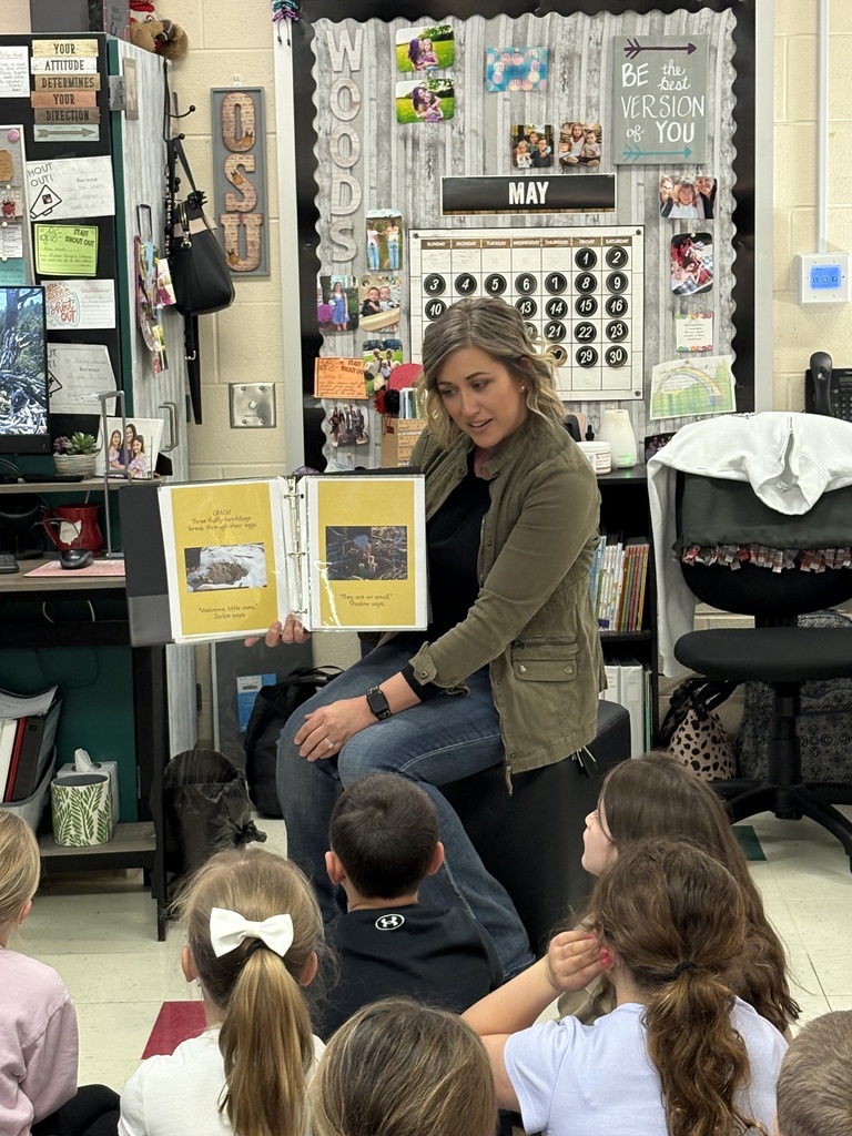 A teacher wearing a green utility jacket and jeans sits before her class, holding open a yellow-paged book that features photos of bald eagles. The page she is showing includes text about hatchlings breaking through their eggs and mentions the eagles Jackie and Shadow. The students, seen from behind, look up attentively while the background shows a classroom wall decorated with photos and motivational signs.