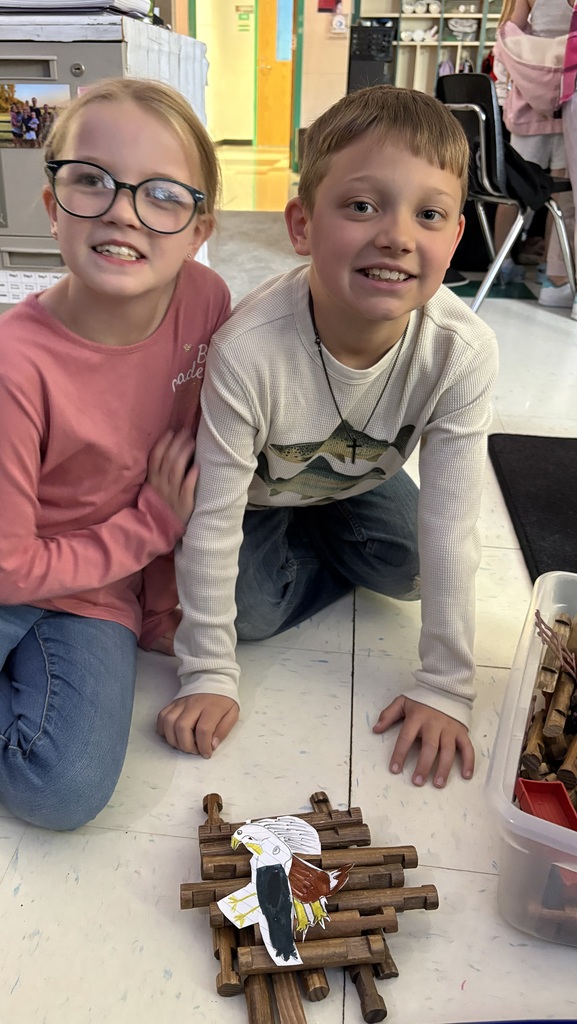 Two smiling children, a girl in a pink shirt and a boy in a white thermal shirt, kneel on a classroom floor next to a creative project. They have constructed a small eagle's nest out of interlocking wooden toy logs and placed a hand-drawn, colored paper cutout of a bald eagle on top. A plastic bin of building toys sits on the floor beside them.