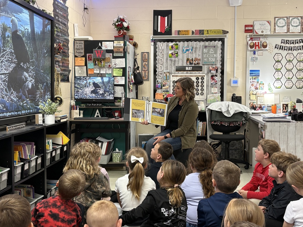 In a brightly decorated elementary classroom, a female teacher sits on a stool and reads a large binder-style book to a group of young students sitting on the floor. To the left, a large monitor and a smaller computer screen both display the live bald eagle nest cam. The room is filled with educational posters, a "Happy Birthday" wall, and a calendar marked for the month of May.