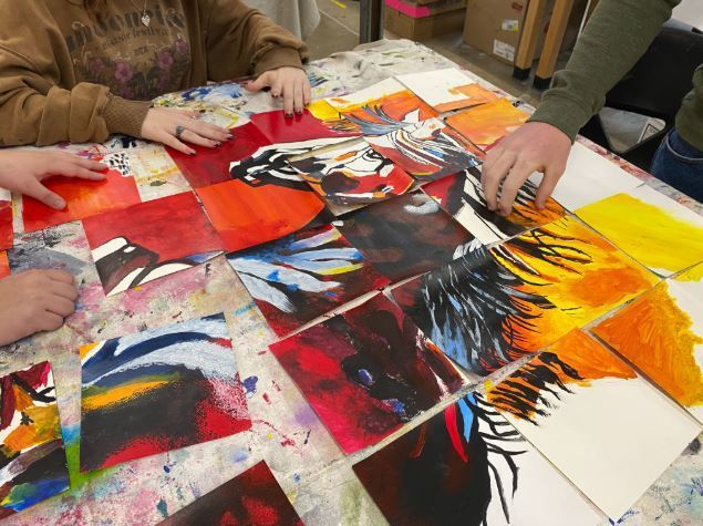 A close-up view shows hands reaching toward the table to align the individual art panels. Each square tile features expressive brushstrokes in vibrant shades of red, orange, yellow, and blue, which, when placed together, begin to form the detailed head and mane of the horse. The table surface beneath is heavily textured with layers of dried paint splatters.