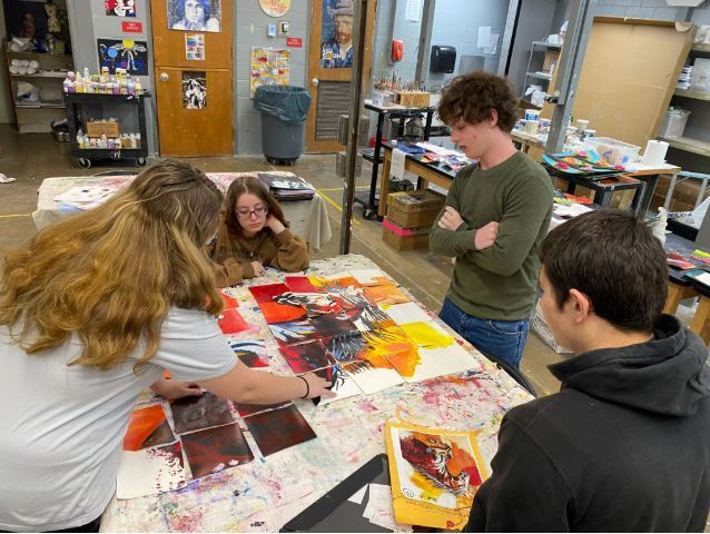 A group of five high school students works together in a cluttered art classroom to assemble a large-scale collaborative painting of a horse. The students are gathered around a paint-stained table, carefully arranging several square painted panels to recreate a reference image of a dynamic, multicolored horse with a flowing mane against a sunset-colored background.