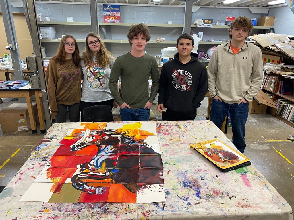 The five students stand behind the finished assembly of their project, posing for a group photo in their art studio. They are dressed casually in hoodies and long-sleeved shirts, looking toward the camera with a sense of accomplishment. The large horse mosaic is laid out completely on the table in front of them, showing the successful transition from individual painted squares to a singular, cohesive piece of art.