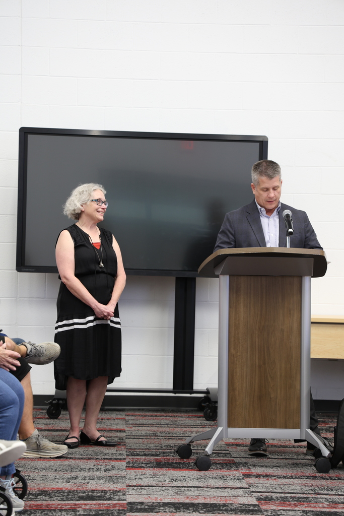 Ms. Amy Fox standing beside Mr. Fife who speaks at a podium in the Community Room