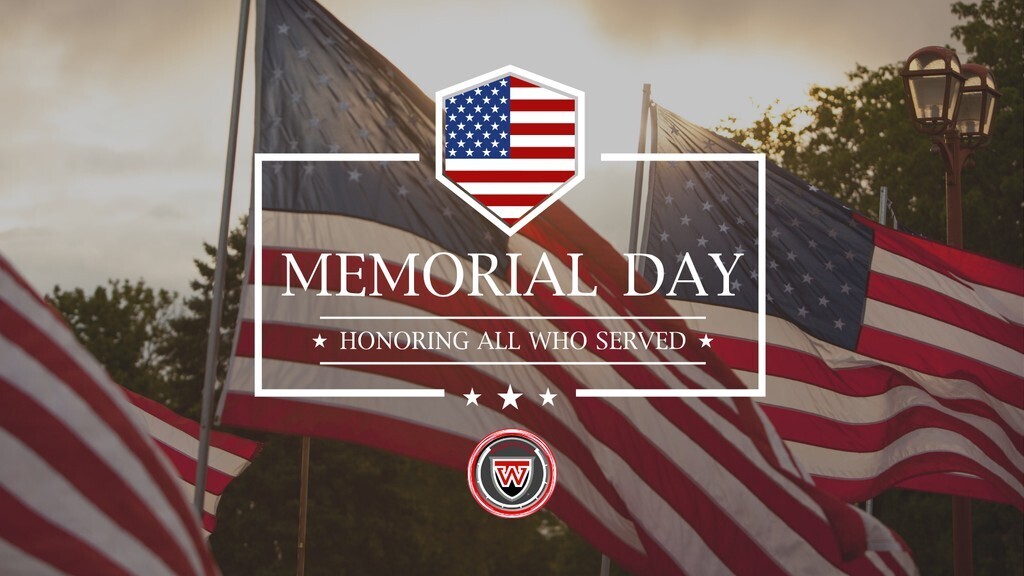 Memorial Day - Graphic featuring the text "MEMORIAL DAY" and "HONORING ALL WHO SERVED" inside a white rectangular border. Above the text is a shield icon containing the American flag, and below it are three small white stars and a circular school logo with a stylized letter "W". The background shows multiple American flags waving outdoors against a soft, overcast sky, flanked by a vintage-style streetlamp.