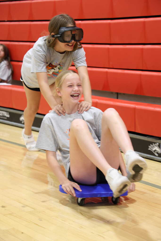 Student sitting on a scooter seat, while another student pushes them while wearing impairment googles