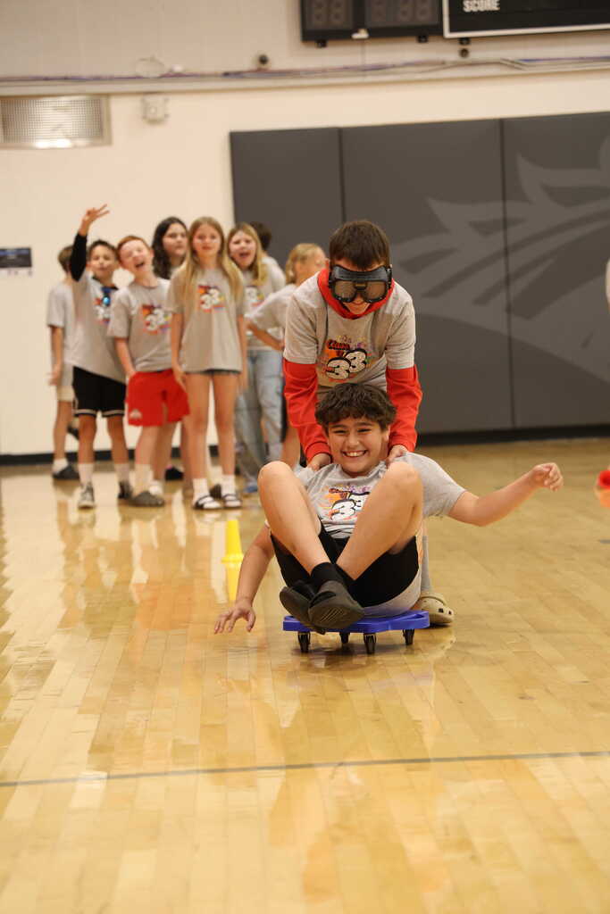 Student sitting on a scooter seat, while another student pushes them while wearing impairment googles
