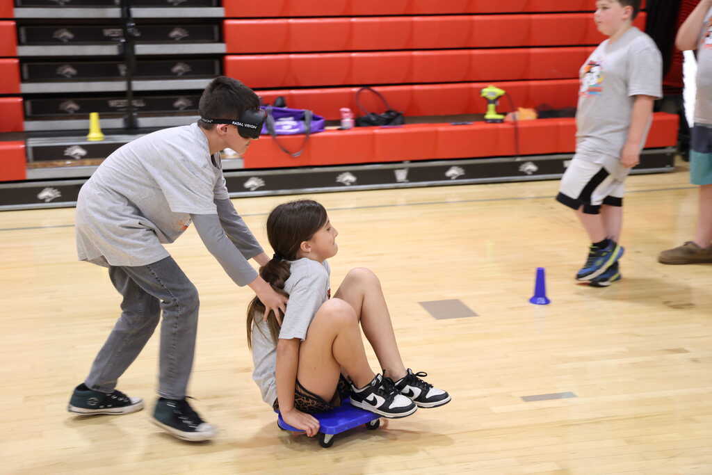 Student sitting on a scooter seat, while another student pushes them while wearing impairment googles