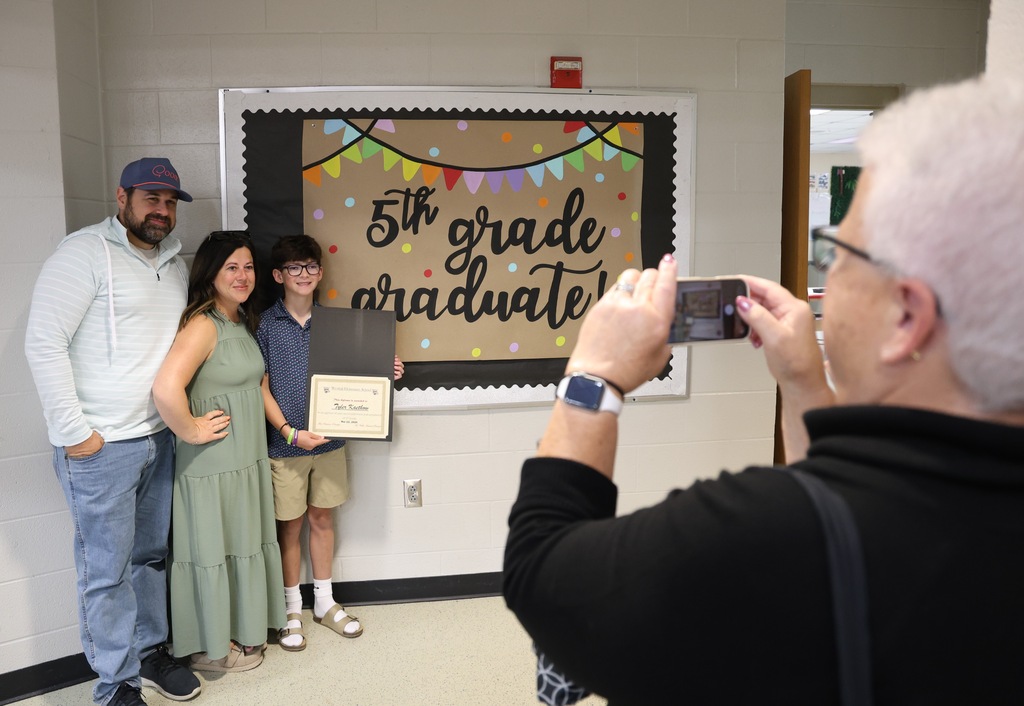 A family posing in front of a sign that reads 5th grade graduate while a female takes a photo on a cell phone