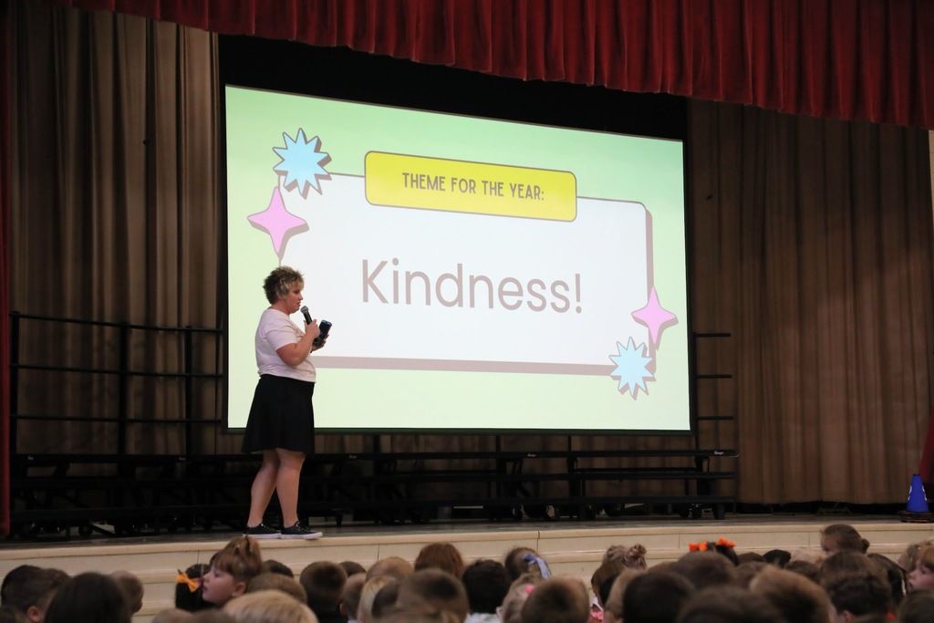 Miss Wynkoop speaking into a microphone in front of a large screen that reads "Theme for the year: Kindness"