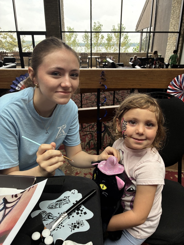 Student and child looking at camera smiling, one is holding a paint brush the other has a spiderweb design painted on her cheek