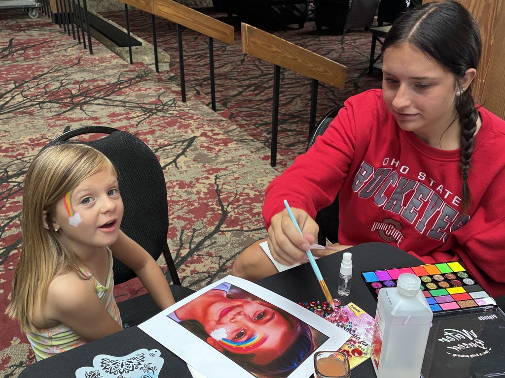 A child smiling at the camera while a student holds a paintbrush
