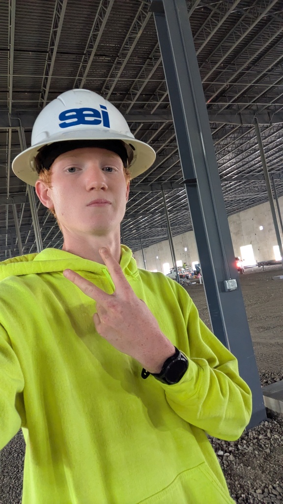 Reid Minor in a bright yellow hoodie and a white hardhat that reads SEI, giving a peace sign to the camera