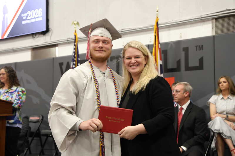 Graduate in light grey cap and gown posing with vocal music teacher Miss McCune while holding a red diploma