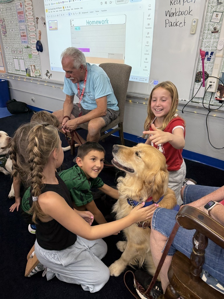 Elementary students happily pet a seated golden retriever therapy dog in a classroom while an adult visitor looks on