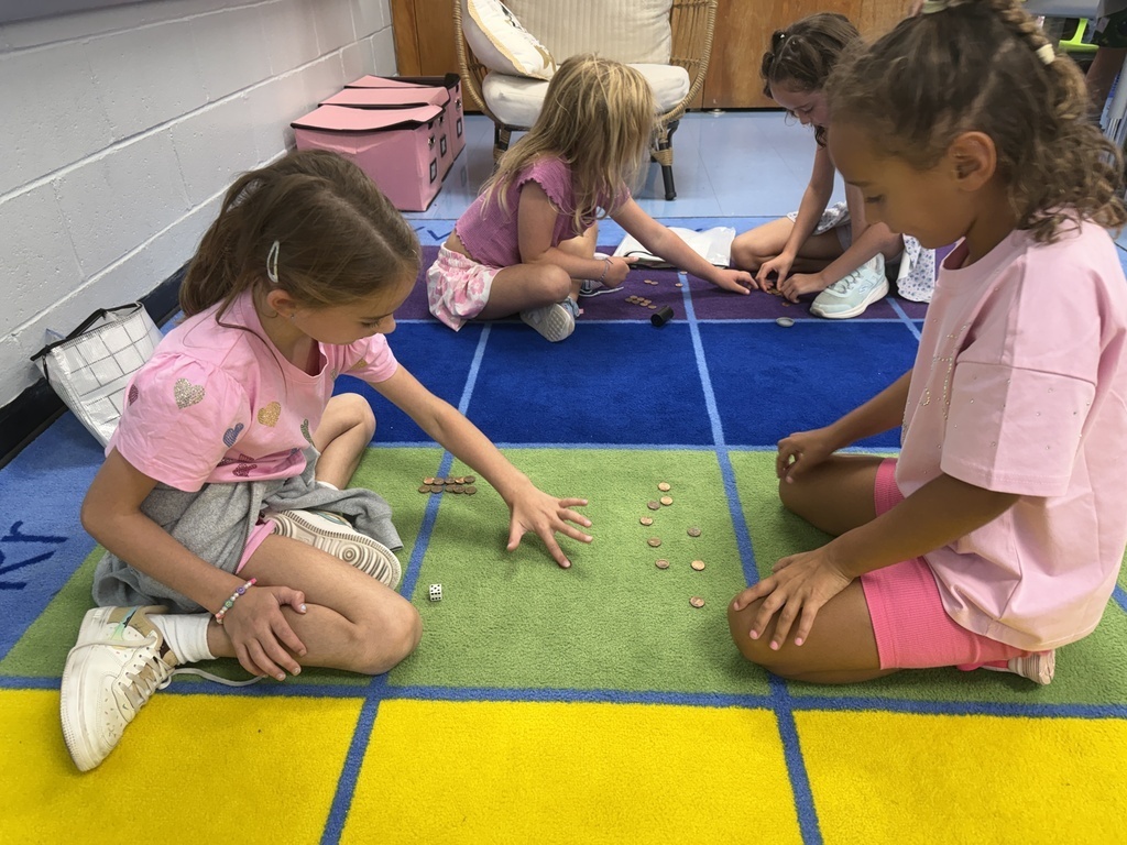 Young students sit cross-legged on a colorful carpet in a classroom, playing a game with coins and dice