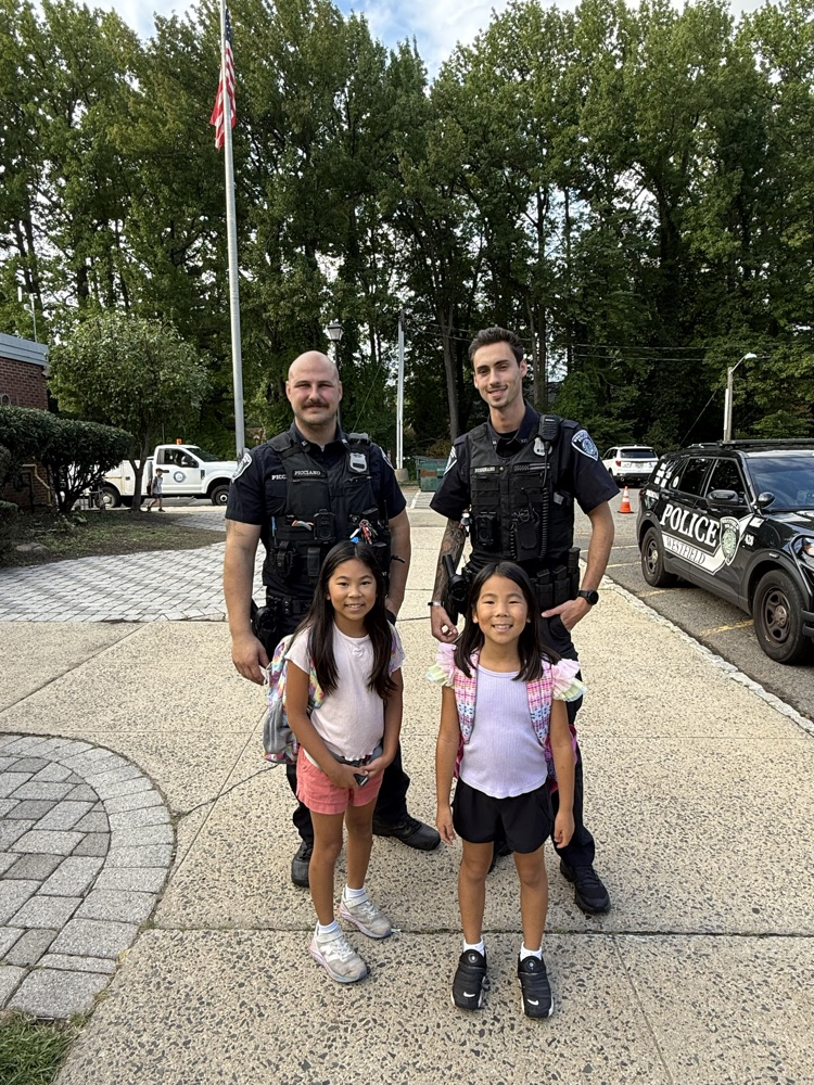 wo smiling young girls stand on a school walkway with two Westfield police officers outside a school building