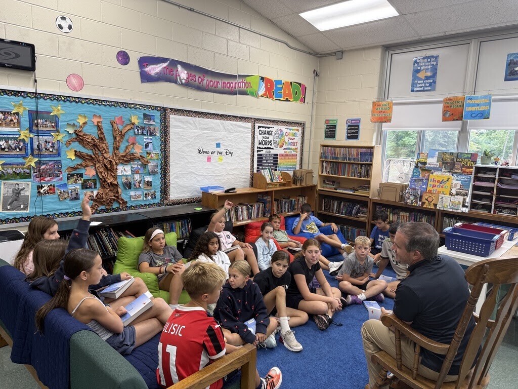 Students sit cross-legged on a blue rug in a classroom, holding up mini whiteboards with their handwritten answers for their teacher, who stands in front of the room.