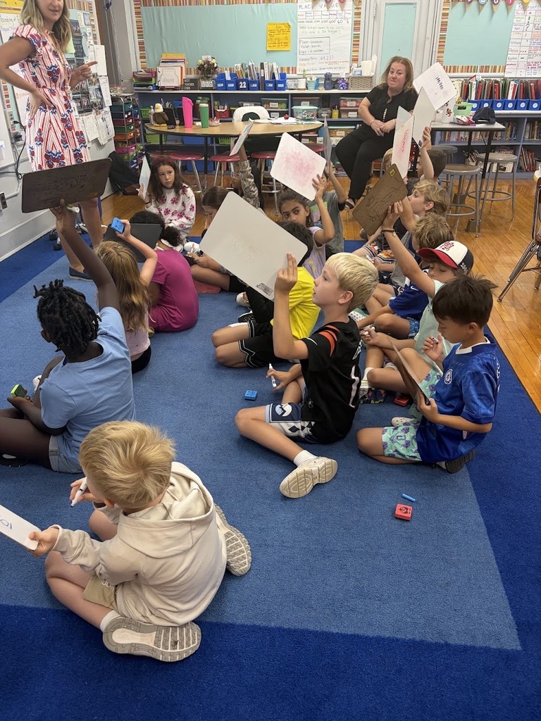 Students sit cross-legged on a blue rug in a classroom, holding up mini whiteboards with their handwritten answers for their teacher, who stands in front of the room.