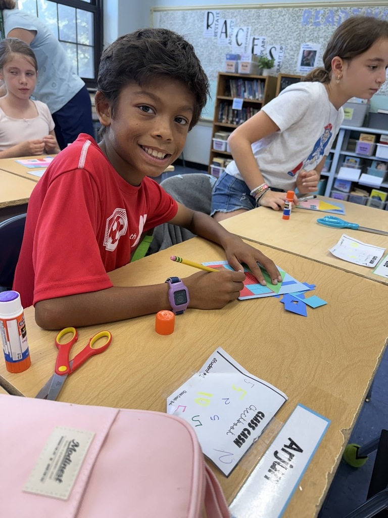 A smiling student wearing a red shirt sits at a classroom desk working on a colorful paper cutout art project, with glue sticks, scissors, and paper shapes spread out on the table.