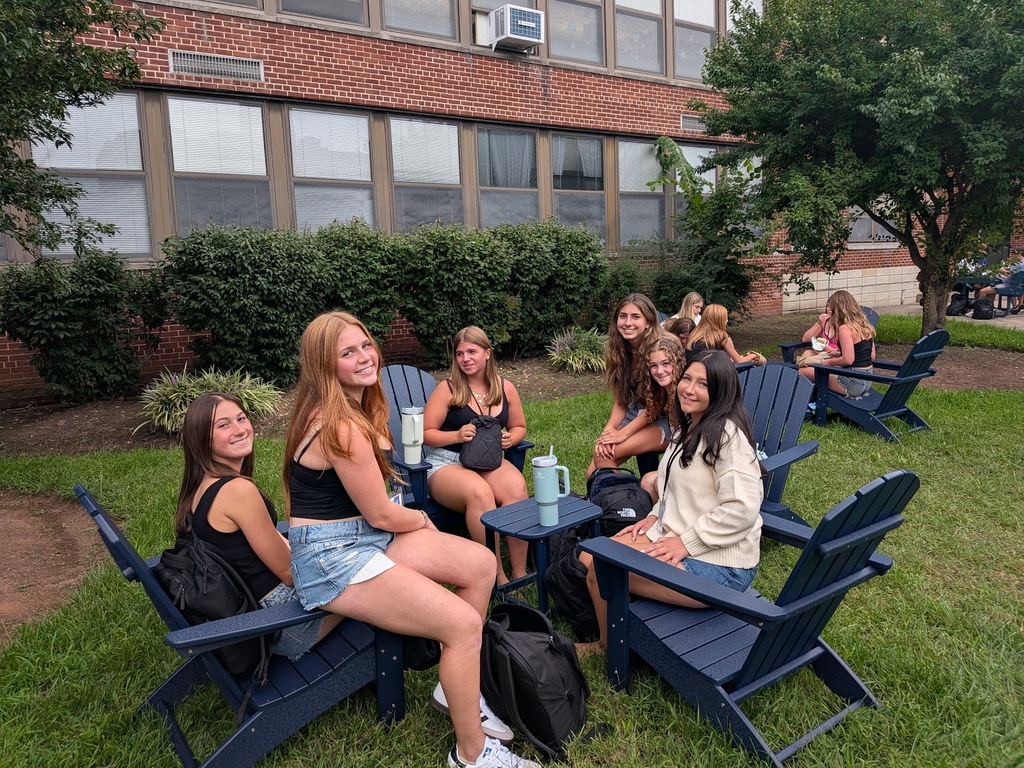 A group of students sits outside in dark blue Adirondack chairs arranged in a circle on a grassy lawn next to a brick school building, smiling and chatting during the school day.