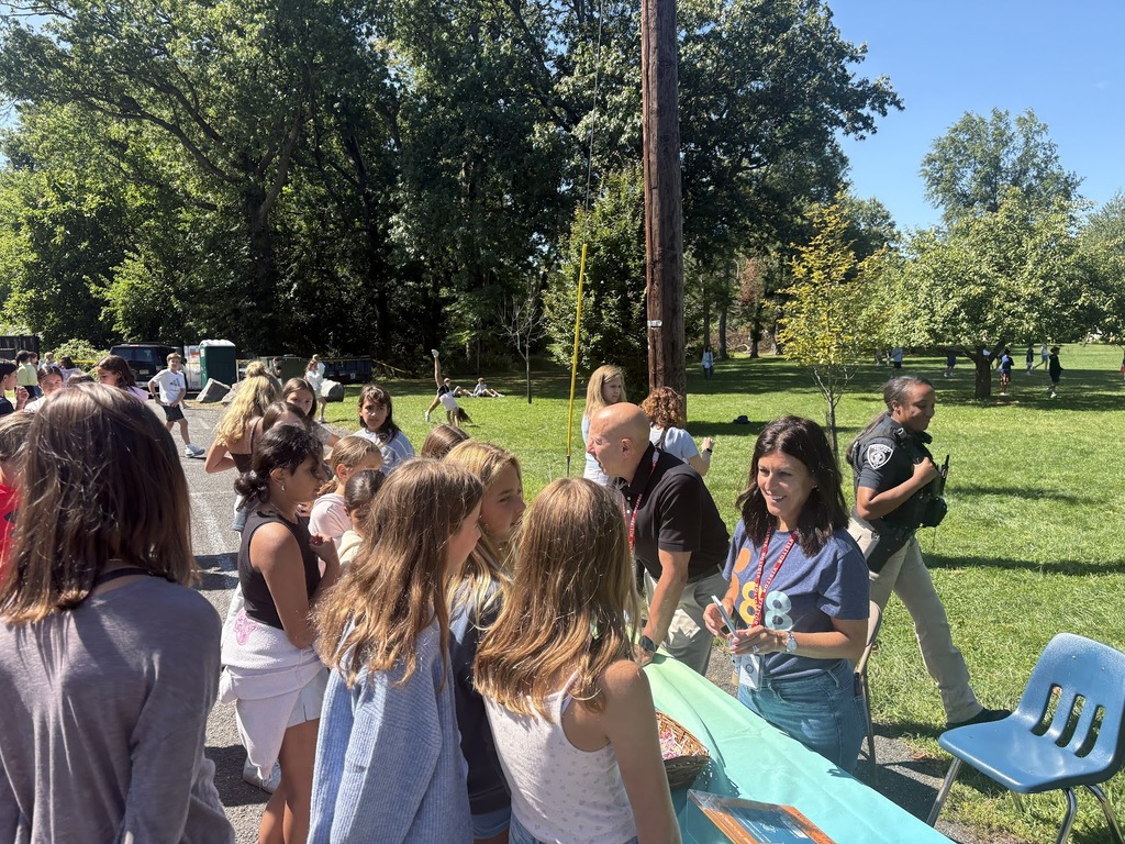 A staff member talks with students standing outdoors in a line near a green table during an outdoor school activity on a sunny day.