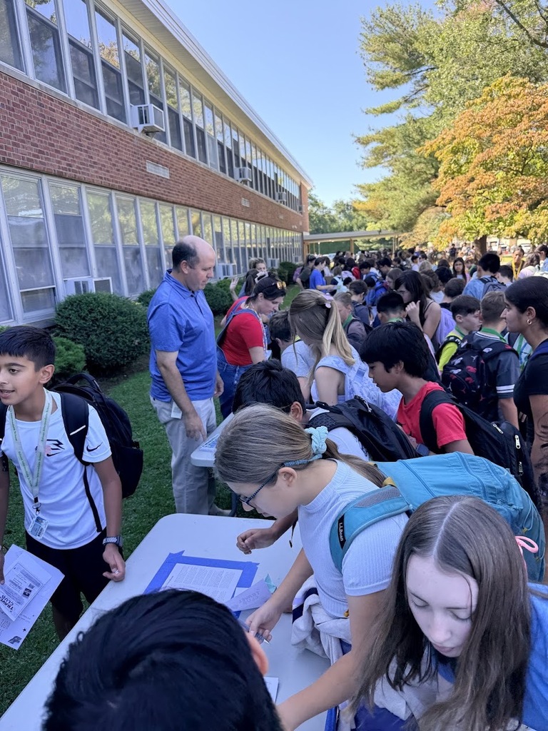 Students gather around outdoor tables set up on the grass near a brick school building while staff members interact with them during a school club fair.
