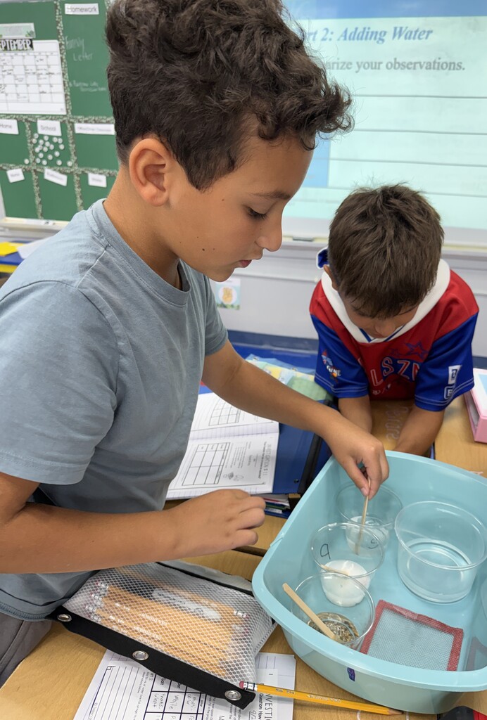 2 students standing at their desks, one student stirring a mixture in a cup, for a science experiment