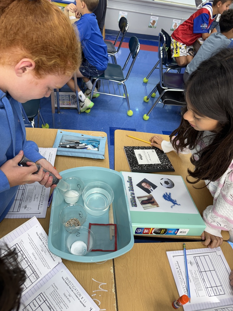2 students standing at their desks, one student squirting water in a cup, for a science experiment
