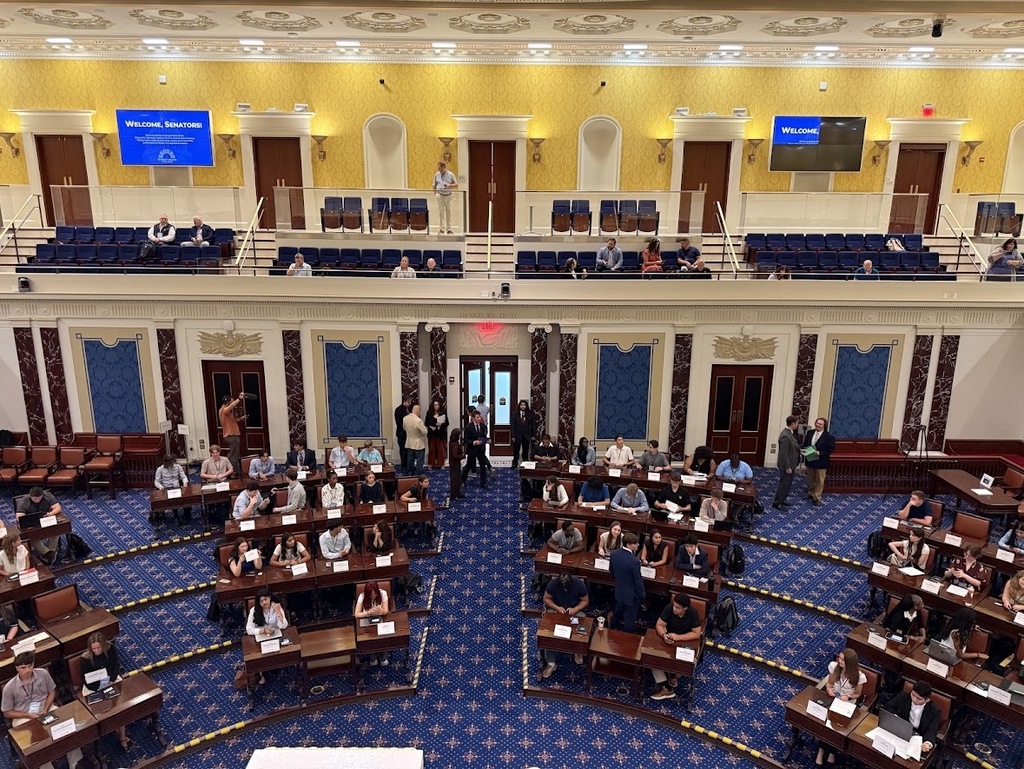 View looking down into a full replica Senate Chamber where student delegates sit at rows of curved wooden desks on blue carpet.