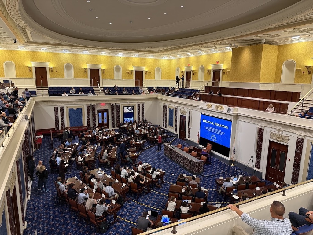 Wide view of student delegates in a replica Senate Chamber with yellow walls and a large screen displaying "WELCOME, SENATORS!"
